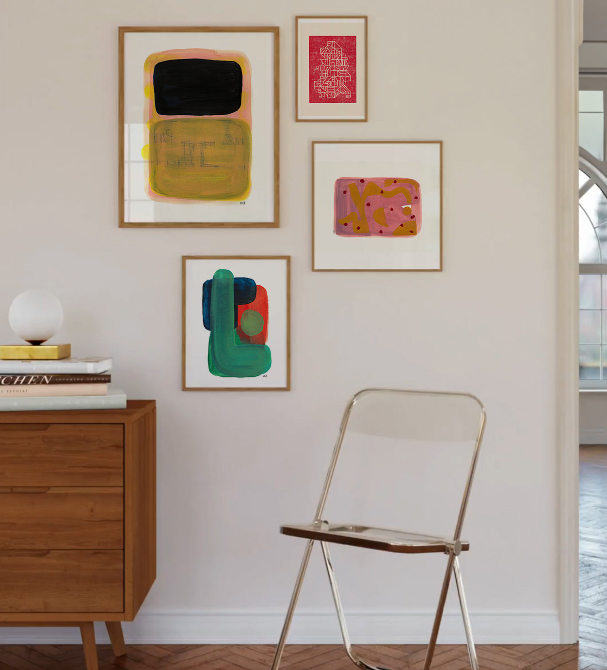 Sunny room with herringbone flooring, showcasing four framed abstract art posters in varying colors and designs. Nearby stands a wooden cabinet with books and a lamp, next to a metal chair."